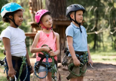 Three kids wearing helmets and harnesses preparing for outdoor ropes course at summer camp.