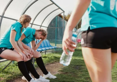 Young soccer player sitting on bench holding knee in pain while teammate offers support on field.